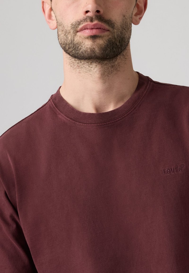 Uomo con barba corta che indossa una semplice maglietta bordeaux Levi's con girocollo, mostrato dal collo fino a metà petto su uno sfondo grigio chiaro.