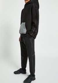 Svart huvtröja med en grå ficka, vit sömnad och justerbara dragsnören, matchad med svarta byxor och svarta sneakers.