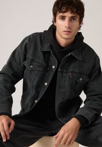 Μαύρο τζιν μπουφάν με ελαφρώς ξεβάμμενη υφή, κλασικός σχεδιασμός, μεταλλικά κουμπιά και δύο τσέπες στο στήθος, φορεμένο πάνω από μια μαύρη κουκούλα.