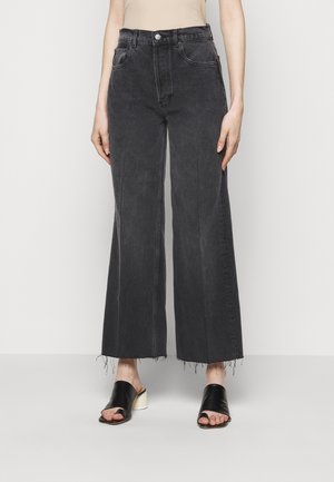 Zwarte jeans met hoge taille, wijde pijpen en rafelige zomen, gedragen met zwarte mulehakken met open tenen en witte blokhakken.