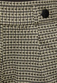 Pantalon à motif pied-de-poule en vert olive et crème, avec un devant plat, un bouton noir et une texture subtile.