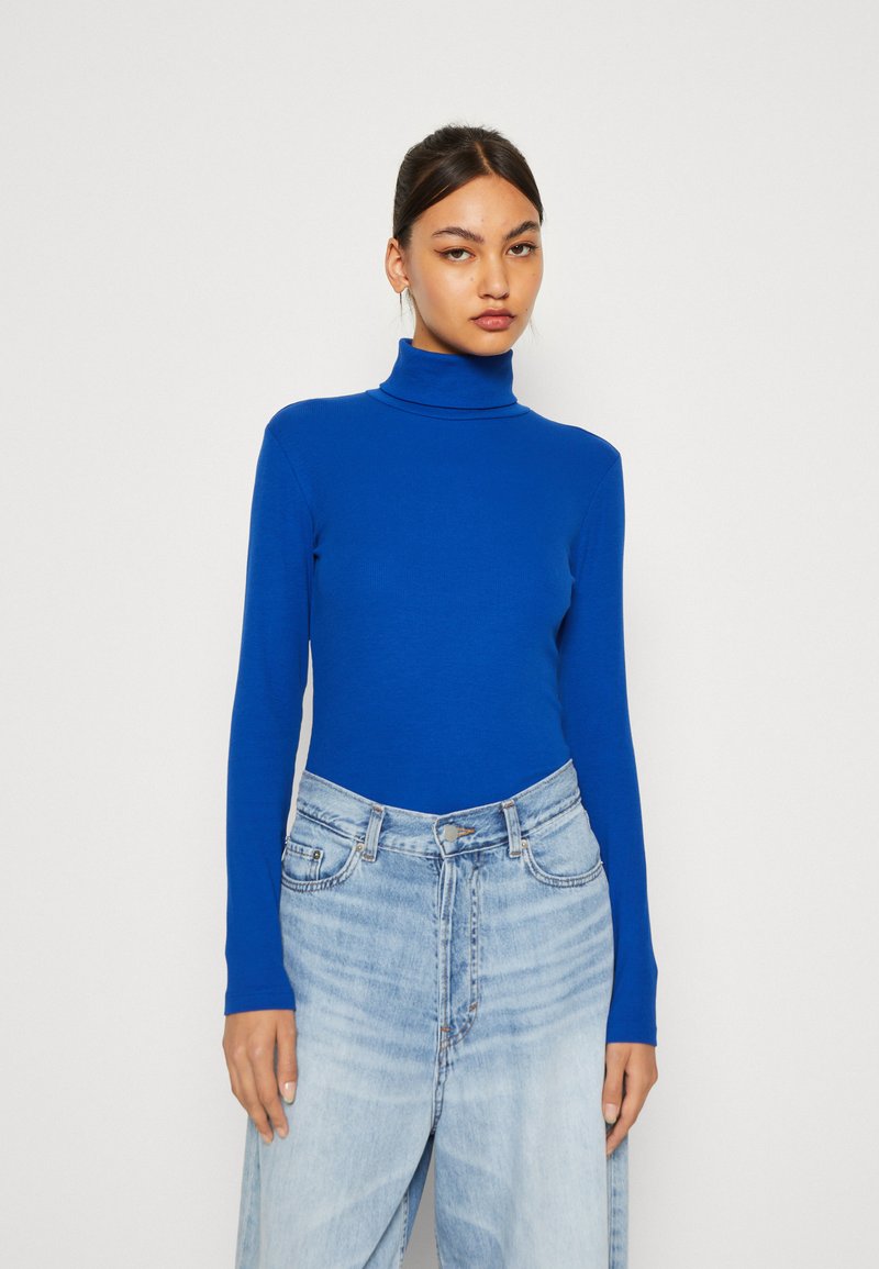Blå høyhalset topp i en tettsittende design, laget av glatt stoff. Kombinert med lyseblå, høyt liv, brede ben jeans med en falmet utseende.