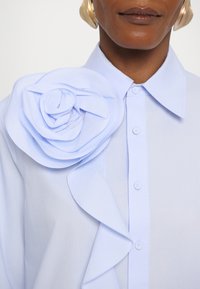 Ανοιχτό μπλε πουκάμισο με εμφανή ύφασμα ροζέτα στον αριστερό ώμο και ρυτιδωτή λεπτομέρεια μπροστά. Τα κουμπιά είναι ορατά κατά μήκος του κέντρου.