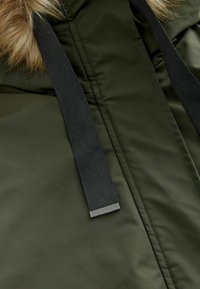 Parka vert olive avec une capuche en fausse fourrure. Présente une sangle en nylon noir et un accent en métal sur la fermeture éclair. Tissu lisse et résistant à l'eau.