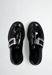 Black patent leather loafers featuring a glossy finish, rectangular rhinestone buckle detail, and rounded toe with a thick sole.