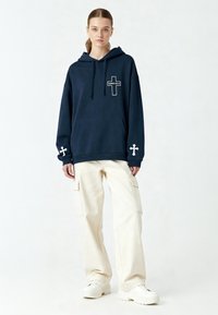 Felpa navy con grafica a croce bianca e testo "KARMA KILLS", dotata di tasca frontale e croci bianche sulle maniche, abbinata a pantaloni cargo color crema.