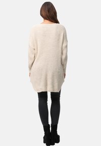 Suéter de punto beige de gran tamaño con mangas largas, textura acanalada y abertura en la parte trasera, combinado con pantalones ajustados negros y botas de tobillo.