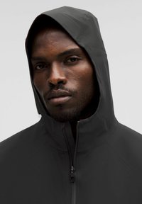 Čierna kapucňová bunda s vysokým golierom a predným zipsom. Vyrobená z hladkej, ľahkej látky, s čistým, minimalistickým dizajnom.