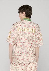 Chemise à manches courtes avec un fond crème orné de motifs de roses rouges et de bouteilles ; détails de texte doré et col rayé vert.