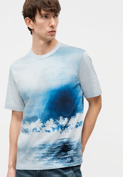 Νέος άνδρας που φοράει μπλε μπλουζάκι με κοντά μανίκια και διαβαθμισμένο χρώμα, με λευκό τύπωμα από φοίνικα και κύμα ωκεανού.