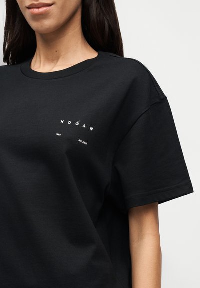 Μαύρο βαμβακερό μπλουζάκι με κοντά μανίκια, με λευκό κείμενο που γράφει "HOGAN 1986 MILANO" στην αριστερή πλευρά του στήθους. Μαλακή, απλή υφή.