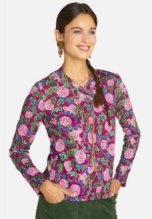 Woman with long braided hair wears a purple floral long-sleeve top, green pants, gold earrings, and a long gold necklace, smiling with hands in pockets.