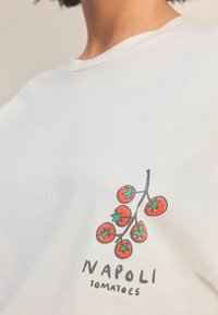 Biela bavlnená tričko s ilustráciou červených paradajok so zelenými listami a čiernym textom "NAPOLI TOMATOES" nižšie.