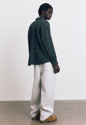 Mlada osoba stoji bočno, nosi tamnozelenu košulju na kockice, svijetlosive široke hlače i smeđe čizme na sivom podu.