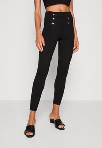 Svarta höga leggings med knappdetaljer fram, slim fit, slät textur och tapered ben-design, kombinerat med svarta öppna klackskor.