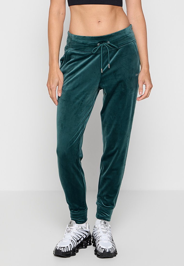 Kobieta w turkusowych dresowych spodniach z weluru z sznurkiem w pasie oraz białych i czarnych sneakerach sportowych, stojąca na tle w jednolitym kolorze.