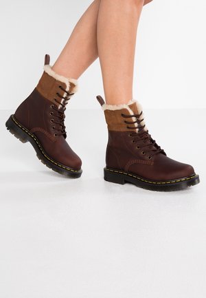 Bottes en cuir marron montant à la cheville, doublées de fourrure, à lacets sur le devant, semelles épaisses noires, avec des coutures jaunes, portées sur des jambes nues.