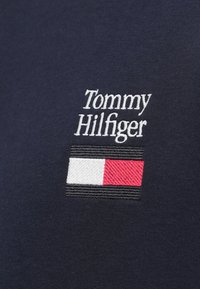 Tommy Hilfiger-logo met witte tekst boven een rechthoekige vlag met witte, rode en zwarte horizontale strepen op marineblauwe stof.
