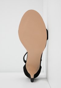 Chaussure à talons hauts en daim noir avec un bout pointu, dotée d'une semelle beige lisse et d'un design minimaliste. Accents clairs sur le talon.
