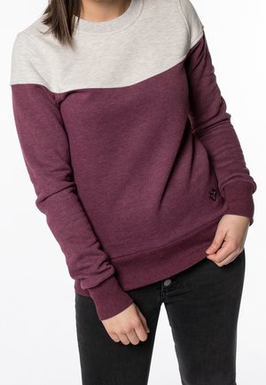 Person wearing a long-sleeve maroon and light gray color-block sweatshirt with black pants, hands relaxed at waist level.