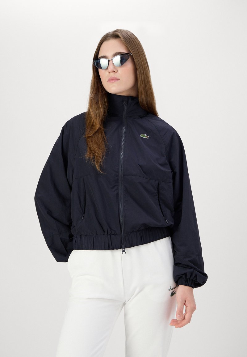 Veste coupe-vent bleue marine courte avec un col haut, un ourlet élastiqué, des poches avant et un logo vert. Associée à un pantalon blanc et des lunettes de soleil.