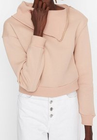 Sudadera corta de color rosa claro con cuello ancho y doblado, puños acanalados y un detalle decorativo de cremallera en el hombro.