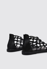 Sandalias negras de punta abierta con diseño trenzado en la parte superior, suela plana y cierre con cremallera en la parte trasera. Fabricadas con material sintético de textura suave.