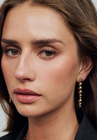 Guldhängande örhängen med flera sfäriska pärlor, slät hud, neutral läppfärg och subtil sminkning mot en mörk bakgrund.