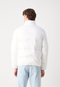 Osoba widziana z tyłu, ubrana w białą kurtkę puchową i jasnoniebieskie dżinsy, na jednolitym jasnym tle.