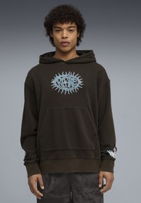 Νεαρός άνδρας με σγουρά μαλλιά που φοράει σκούρο φούτερ με κουκούλα με λογότυπο "RARE" σε σχήμα ήλιου και παντελόνι με σχέδια, στέκεται μπροστά σε γκρι φόντο.