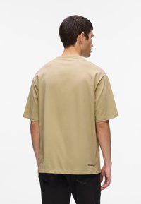 Olive žalia medvilnės crewneck T-shirt, laisvas kirpimas, trumpomis rankovėmis, su maža logo netoli apatinės nugaros dalies. Minkšta tekstūra.