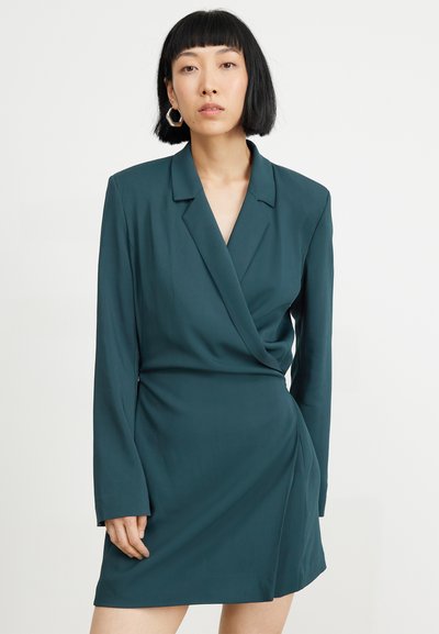 Tiefgrünes Wickelkleid mit langen Ärmeln, einem eingeschnittenen Kragen und einer drapierten Vorderseite. Hergestellt aus glattem Stoff mit einer leicht strukturierten Silhouette.