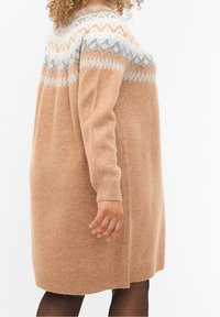 Robe pull oversize en beige clair, avec un empiècement à motifs en crème, gris et pêche. Manches longues et poignets côtelés. Texture douce.