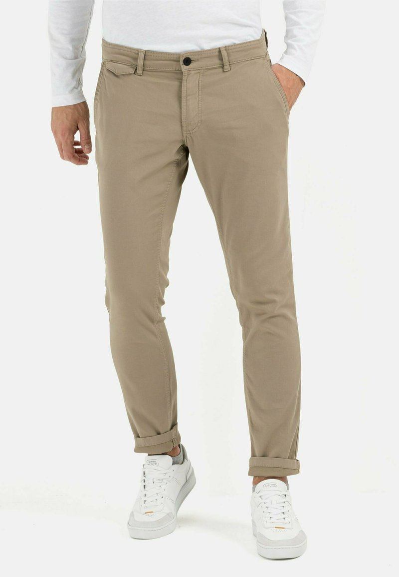Homme portant un pantalon beige slim-fit retroussé aux chevilles, une chemise blanche à manches longues et des baskets blanches, debout avec une main dans la poche.