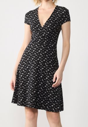 Black short-sleeve dress with white floral print, V-neckline, fitted waist, and flared skirt worn by a standing person.