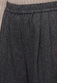 Gros plan sur un pantalon en tissu gris foncé texturé avec une taille élastique et des plis verticaux, porté avec un pull beige côtelé.