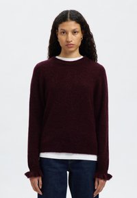 Vino rdeč pulover iz mehke, teksturirane tkanine, z okroglim izrezom in strganimi manšetami. Oblečen preko bele srajce in kombiniran s temnimi kavbojkami.