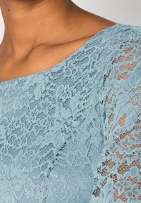 Top en dentelle bleu clair avec un motif floral, des coutures délicates et des manches longues transparentes. L'encolure est large et arrondie.