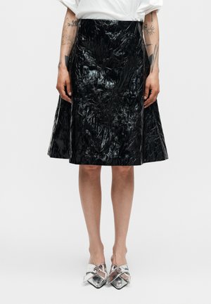 Kvinde iført en skinnende sort tekstureret knælængde nederdel, hvid bluse, sølvfarvede spidse sko, med synlige tatoverede arme.