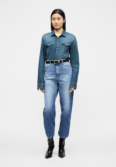 Γυναίκα που φοράει σκούρο πουκάμισο από τζιν με κουμπιά και τσέπες στο στήθος, ανοιχτόχρωμο τζιν ψηλόμεσο, μαύρη ζώνη και μαύρα μποτάκια μέχρι τον αστράγαλο, στέκεται μπροστά σε λευκό φόντο.