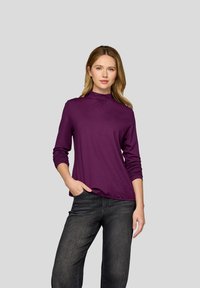 Frau mit hellbraunen Haaren, die ein langärmliges lila Oberteil mit Stehkragen und schwarze Jeans trägt, steht mit einer Hand in der Tasche vor einem grauen Hintergrund.