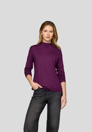 Woman with light brown hair wearing a long-sleeve purple mock-neck top and black jeans, standing with one hand in pocket against gray background.