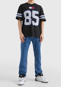 Černé tričko s krátkým rukávem se velkým bílým číslem 85, modrými akcenty a logem Tommy Jeans. Odpovídá s světle modrými rovnými džíny a teniskami.