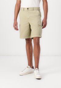 Khaki cargo shorts made of cotton with side pockets. Features a button closure, belt loops, and a straight cut. Paired with white sneakers.