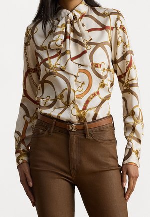 Vrouw draagt een crèmekleurige blouse met lange mouwen met een bruin en goudkleurig ketting- en riemprint, gecombineerd met een bruine broek en een bruine riem met RL gesp.