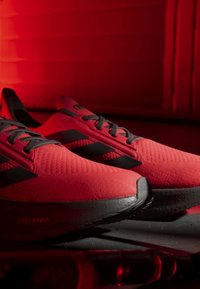 Chaussures de course Adidas rouges et noires avec une semelle Light Boost, placées sur une surface sombre sur fond rouge.