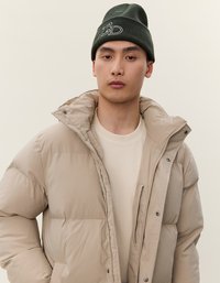 Giovane uomo con espressione neutra che indossa un piumino beige sopra una camicia crema e un berretto verde scuro su uno sfondo chiaro e semplice.