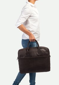Dunkelbraune Lederaktentasche mit doppelten Griffen, einer vorderen Reißverschluss-Tasche und einem Gepäckanhänger, die eine glatte Oberfläche und ein strukturiertes Design aufweist.
