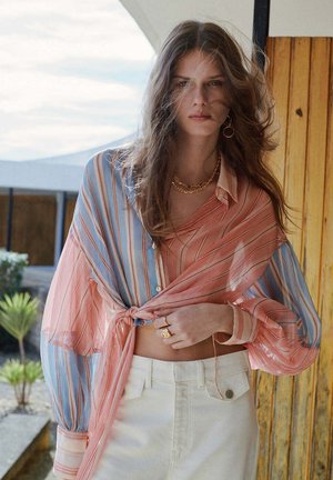 Frau mit lockerem welligem Haar trägt eine gebundene, gestreifte Bluse in Pfirsich- und Blautönen und hoch taillierte weiße Hosen, steht draußen neben einer Holzverkleidung.