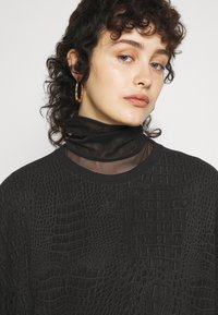 Sweatshirt noir avec un motif crocodile texturé, présentant une couche translucide à col montant en dessous, et un petit logo Adidas sur le devant.